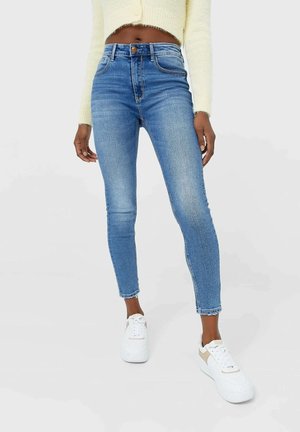 Skinny blue denim jeans featuring a high waist, light fading, and raw hem. Paired with white sneakers and a cropped light yellow sweater.