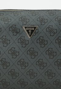 Musta kuvioitu synteettinen clutch, jossa on kolmionmuotoinen metallilogo. Varustettu kohokuvioidulla designilla ja vetoketjukiinnityksellä yläreunassa.