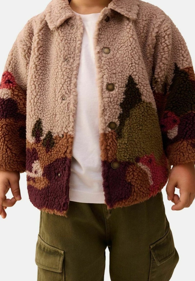 Enfant portant une veste polaire colorée, boutonnée et texturée par-dessus une chemise blanche et un pantalon cargo vert olive.