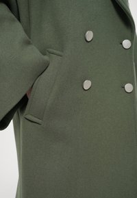 Green wool coat featuring a double-breasted design, large silver buttons, and a side pocket. Smooth texture and structured fit.