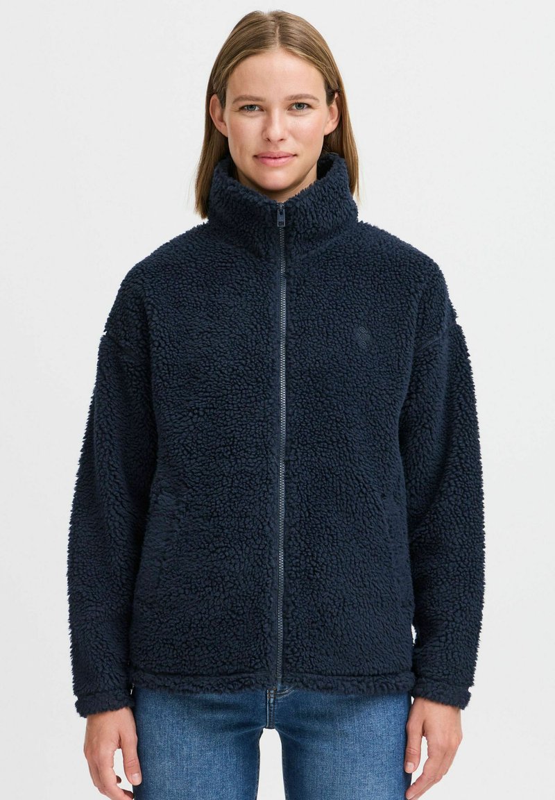 Veste en polaire marine avec col haut et fermeture éclair. Présente un tissu texturé et doux, ainsi que des épaules tombantes. Le modèle porte un jean bleu.