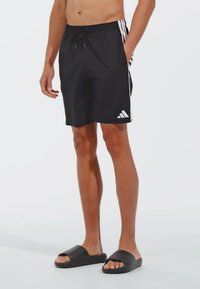 Crne kupaće gaće s bijelim bočnim trakama i elastičnim pojasom s vezicom. Uparene s crnim kliznim sandalama. Mekani, lagani materijal.