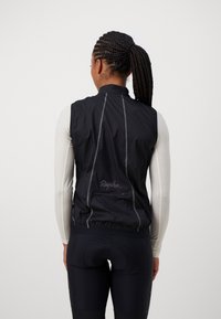 Schwarze Radfahren-Weste mit hohem Kragen, reflektierenden Akzenten, einer Reißverschlusstasche und elastischem Saum. Tragen über einem hellen Langarmoberteil.