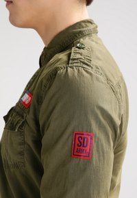 Camicia verde oliva con colletto abbottonato, toppa sul braccio sinistro con "SD" e "ARMY" in rosso e blu, tessuto testurizzato, design militare.