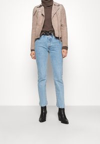 Béžová semišová motorkářská bunda se stříbrnými zipy, hnědý rolák, světle modré džíny přímo střižené a černé kotníkové boty na podpatku.