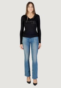 Maglione aderente nero a maniche lunghe con scollo a V e logo embellito, abbinato a jeans blu a zampa d'elefante e scarpe nere con tacco alto.