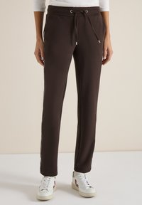 Personne portant un pantalon à cordon de serrage marron foncé et des baskets blanches, debout devant un fond neutre.