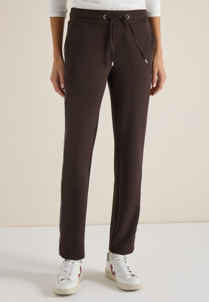 Personne portant un pantalon à cordon de serrage marron foncé et des baskets blanches, debout devant un fond neutre.