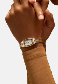 Montre argentée et dorée avec un boîtier rectangulaire, des accents dorés et une lunette sertie de cristaux, portée sur un poignet avec une chemise à manches longues marron.