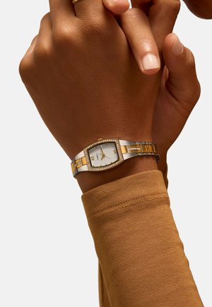 Montre argentée et dorée avec un boîtier rectangulaire, des accents dorés et une lunette sertie de cristaux, portée sur un poignet avec une chemise à manches longues marron.