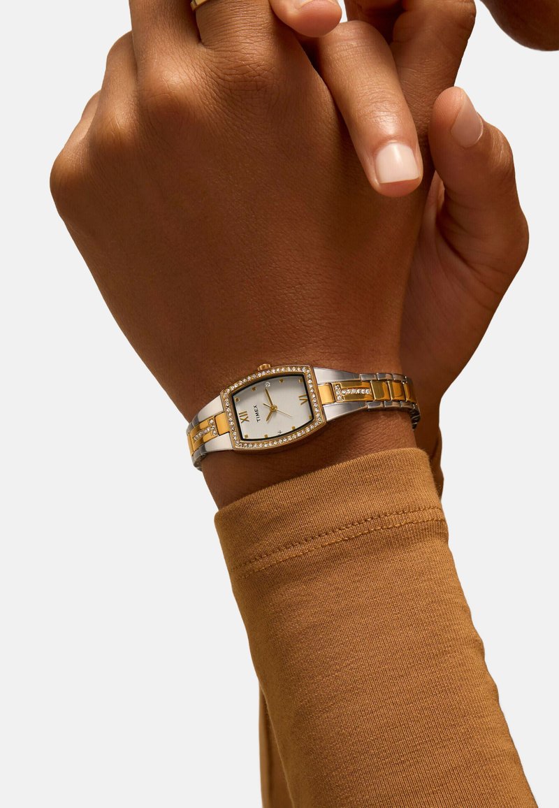 Montre argentée et dorée avec un boîtier rectangulaire, des accents dorés et une lunette sertie de cristaux, portée sur un poignet avec une chemise à manches longues marron.