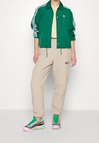 Chaqueta verde de poliéster con rayas blancas, pantalones cargo beige con detalles en los bolsillos y zapatillas deportivas multicolores con acentos en rosa y verde.