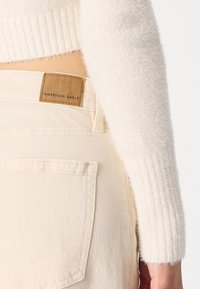 Jeans beige chiaro con un'etichetta in pelle marrone chiaro che recita "American Eagle" sulla cintura; un maglione di peluche crema parzialmente visibile.