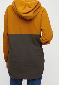 Hooded fleecejack met een mosterdgele bovenkant en een donkergrijze onderkant, met een ontspannen pasvorm en een zachte textuur.