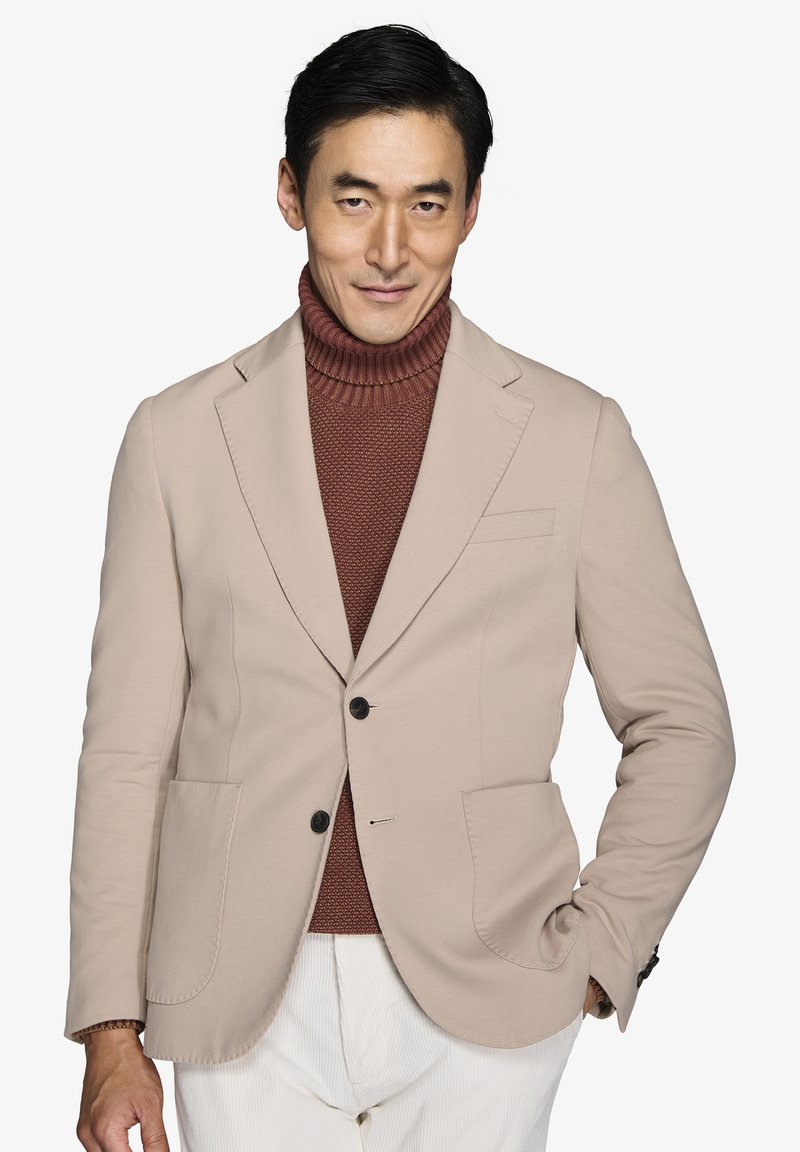 Beige blazer med en skræddersyet pasform, to forlommer og sorte knapper, båret over en rustfarvet turtleneck i en struktureret strik.