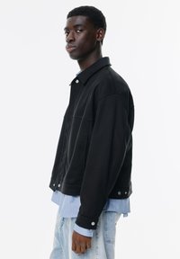 Jachetă din denim negru cu guler, buzunare frontale și nasturi tip capse. Purtați-o peste o cămașă cu dungi albastre deschise și asociați-o cu blugi din denim deschis.