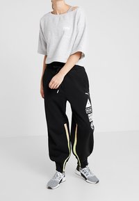 Grijs cropped t-shirt met een klein logo, gecombineerd met zwarte joggers met witte graphics en gele en roze zijpanelen. Grijze sneakers.