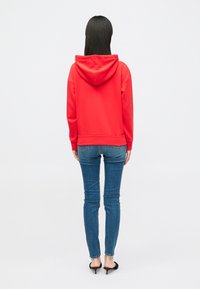 Raudona gobtuvė su atsipalaidavusiu siluetu, derinama su lieknais mėlynais džinsais. Gobtuvė turi raištelį ir iškilnius rankogalius.