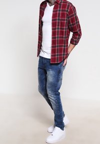 Camisa de cuadros roja y negra sobre una camiseta blanca, combinada con jeans azules desgastados y zapatillas blancas. Atuendo casual con texturas variadas.