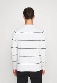 Maglione bianco a maniche lunghe con strisce orizzontali nere, polsini e orlo a coste, con scollo rotondo. Indossato con pantaloni neri.