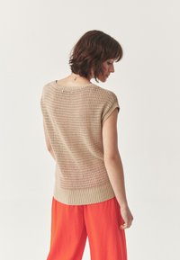 Maglione beige a maglia con maniche corte e motivo a trama aperta, abbinato a pantaloni larghi arancioni. Design testurizzato con orlo a coste.