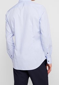 Vaaleansininen pitkähihainen paita, jossa on hillitty ruutukuosi. Paidassa on teräväkauluksinen, nappihihnat ja kaarrettu helma. Sileä puuvillakangas.