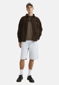 Camisa de corduroy marrón, camiseta verde oliva, pantalones cortos de mezclilla azul claro, zapatos negros y calcetines blancos. Conjunto casual con un corte relajado.