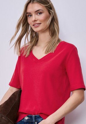 Młoda kobieta z blond włosami ma na sobie czerwoną koszulkę z krótkim rękawem i dekoltem w serek oraz trzyma ciemnobrązową zamszową kurtkę na tle gładkiego jasnego tła.