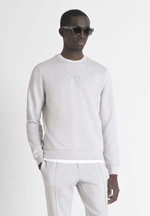 Sudadera gris claro con cuello redondo y puños acanalados, que presenta un logotipo en relieve en la parte frontal. Combinada con pantalones gris claro a juego.