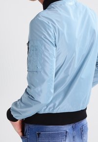 Světle modrá bomber bunda vyrobená z lesklého nylonu s černým žebrovaným límcem a manžetami, která má zapínací kapsu na rukávu a elastický pas.