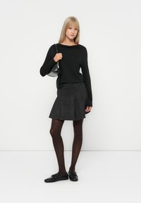 Haut noir à manches longues associé à une jupe à carreaux gris avec des plis. Le modèle porte des collants sombres et des chaussures noires, tenant un petit sac.