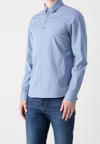 Uomo che indossa una camicia azzurro chiaro a maniche lunghe con un piccolo logo "ck" sul petto, abbinata a jeans blu.