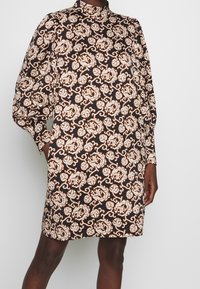 Vestido marrón con un estampado floral en color crema y dorado. Presenta un cuello alto y mangas largas, con un corte recto y bolsillos laterales.