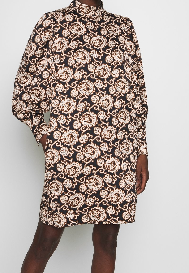 Braunes Kleid mit einem floralen Muster in Creme und Gold. Es hat einen hohen Kragen und lange Ärmel, mit einem geraden Schnitt und Seitentaschen.