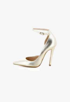 Une chaussure à talon haut dorée brillante avec bout pointu et bride à la cheville avec boucle, présentée sur fond blanc.