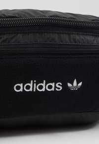 Černý pouzdro s texturovaným fleecovým povrchem, vyznačující se bílým logem "adidas" a logem tří pruhů; zahrnuje černý zip na vrchu.