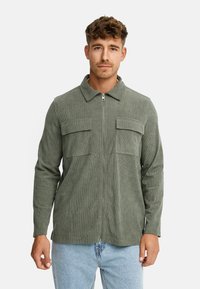 Mand iført en olivengrøn corduroy zip-up jakke med to brystlommer og lyseblå jeans, stående foran en ensfarvet baggrund.