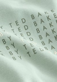 Helles grünes Gewebe mit wiederholten Textmustern in dunklerem Grün und Weiß. Das Material wirkt weich und hat eine feine gerippte Textur.