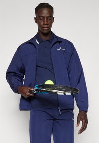 Marineblå vindjakke med elastiske mansjetter, matchende marineblå piké og bukser. Holder en tennisracket med en gul tennisball foran.