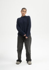 Navy blaues, geripptes Strickpullover mit hohem Kragen und langen Ärmeln, kombiniert mit dunkelgrauen Hosen und hellgrauen Slippern.