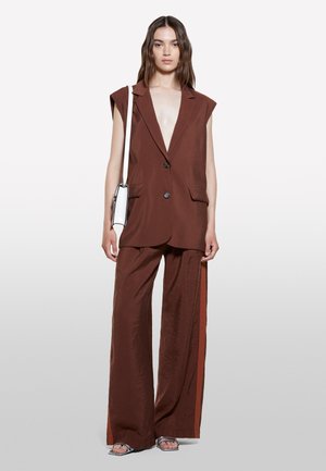 Donna che indossa un blazer marrone senza maniche e pantaloni a gamba larga con strisce laterali arancioni, porta una borsa a tracolla bianca e indossa sandali aperti.