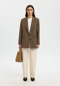 Žena oblečená v hnědém saku, bílé košili, krémových kalhotách a tmavých balerínách, držící světle hnědou semišovou kabelku, stojí na jednobarevném světlém pozadí.