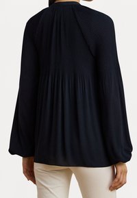 Kvinna klädd i en svart långärmad plisserad blus med lösa ärmslut och vita byxor, sedd bakifrån.