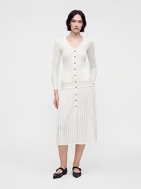 Femme portant une robe blanche texturée à manches longues boutonnée, avec deux poches frontales et des chaussures plates noires à brides à la cheville.