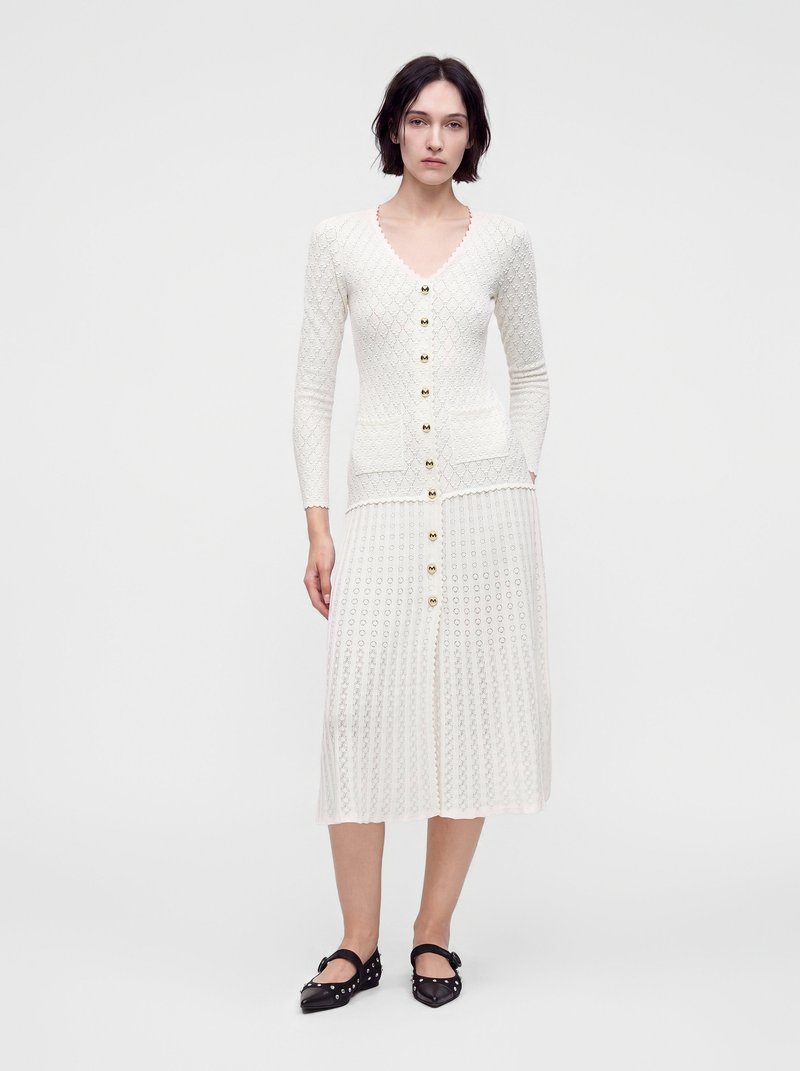 Femme portant une robe blanche texturée à manches longues boutonnée, avec deux poches frontales et des chaussures plates noires à brides à la cheville.