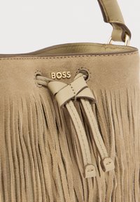 Τσάντα χειρός από μπεζ σουέντ με διακοσμητικές φουντίτσες, φιόγκο στολισμένο και χρυσό λογότυπο στην κορυφή. Ολοκληρώνεται με λείες ιμάντες ως αξεσουάρ.