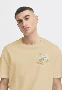 Beige cotton T-shirt featuring a graphic design on the chest with a lemon, newspaper, and pastry illustrations. Short sleeves, crew neck.