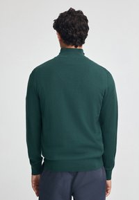 Grön stickad tröja med hög krage, långa ärmar och ribbad fåll. Slät textur, figurnära passform och inga synliga mönster eller detaljer.