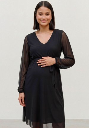 Une femme enceinte souriante se tient devant un mur uni, une main posée sur son ventre. Elle porte une robe noire aux manches transparentes, nouée à la taille.