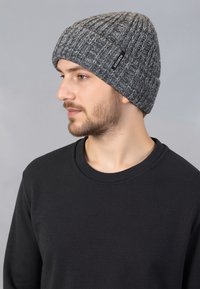Grijze gebreide beanie met een ribbelstructuur en een klein zwart label. Draagt een zwart longsleeve shirt, weergegeven met een close-up profielweergave.